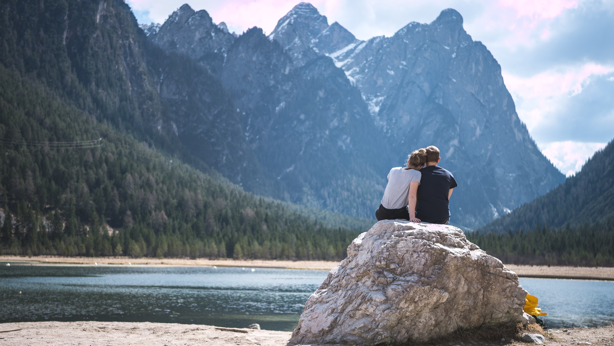 A couple sits with their backs to us and their heads resting together as they look at a beautiful lake and mountain.