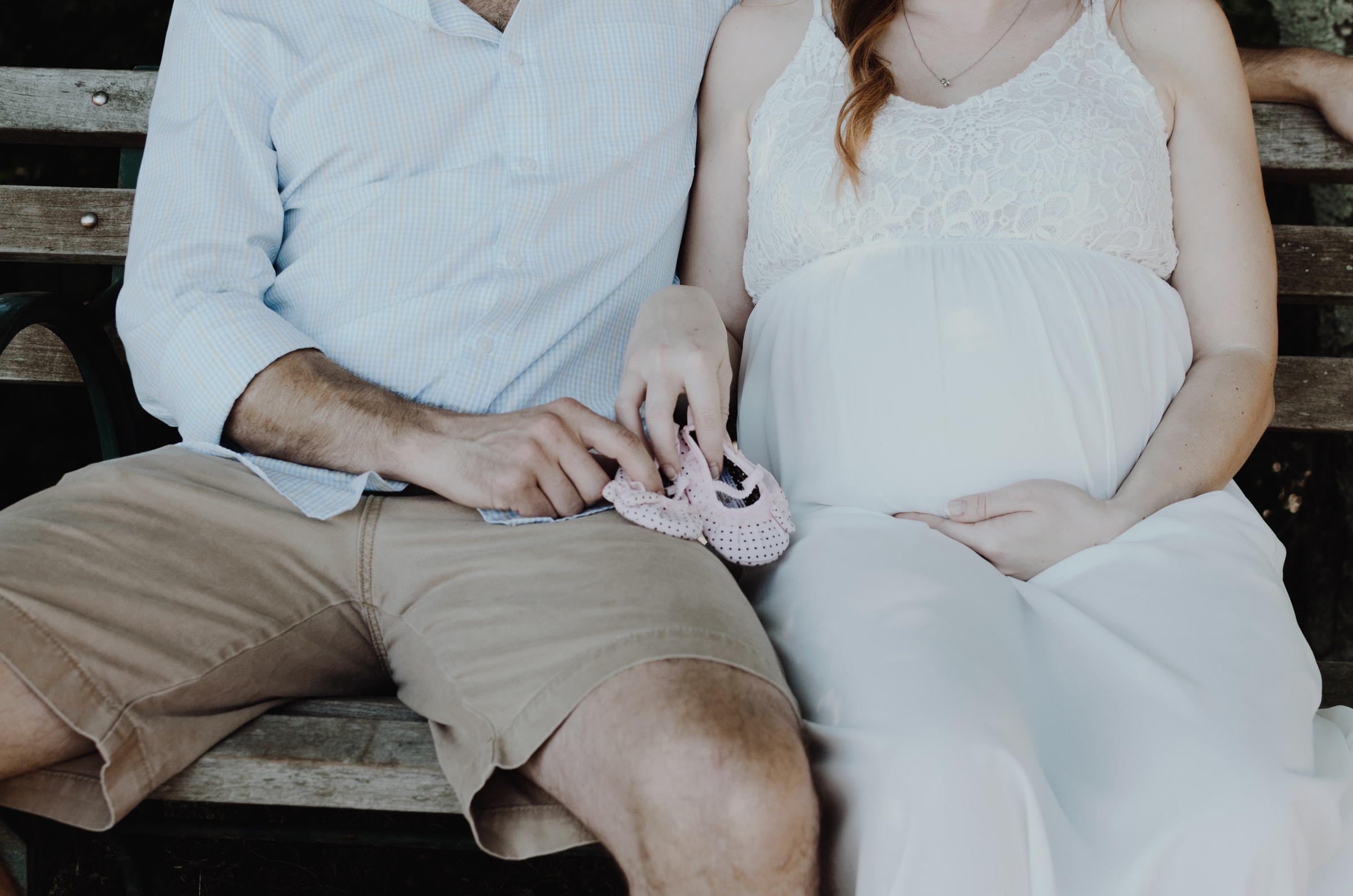 A couple sits on a bench, both holding a pair of pink baby shoes. The woman is pregnant and the man's arm is wrapped around her.