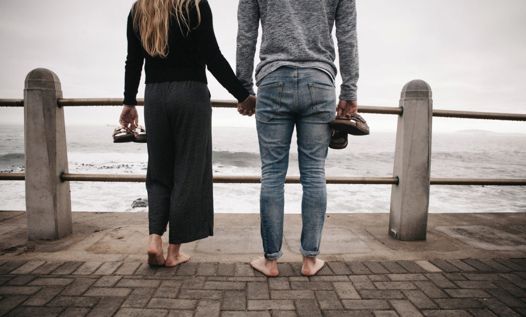 A couple stands with their shoes off, holding hands and looking out at the ocean.