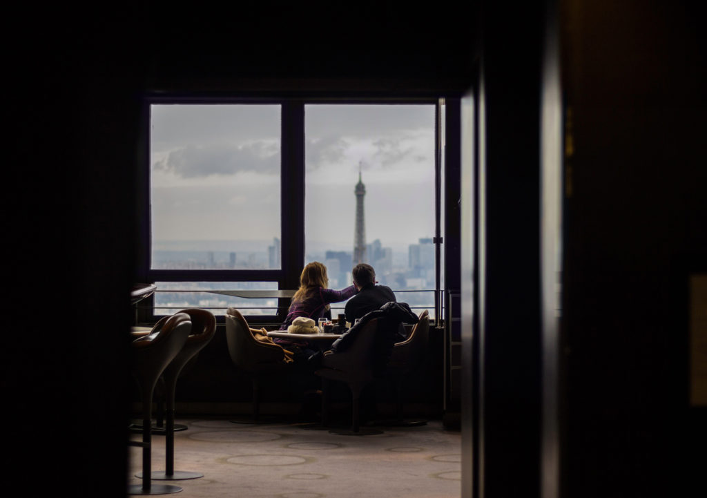 A couple sit at a restaurant table overlooking a city, pointing in the distance.