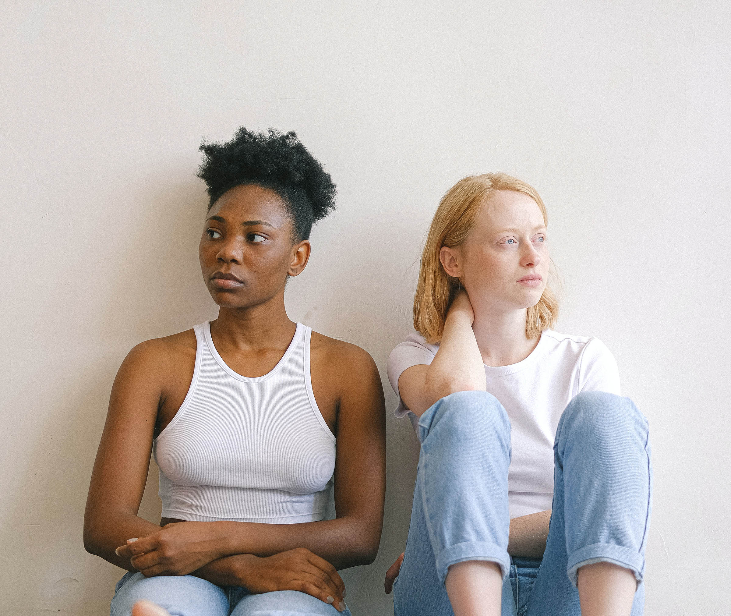 Two women sit against a wall, looking away from each other.