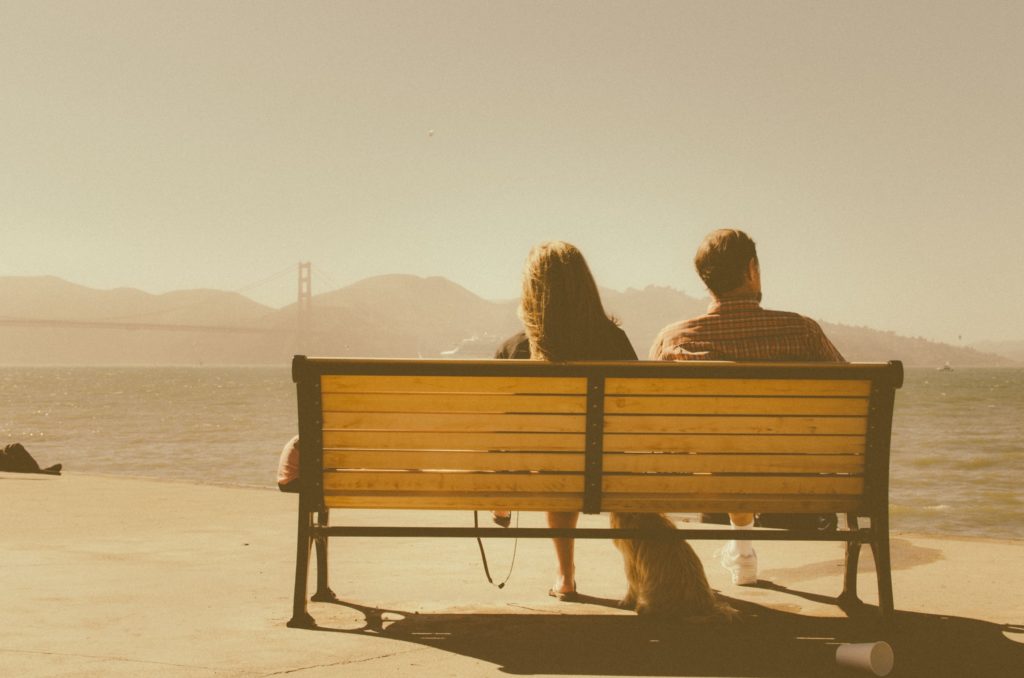 A man and a woman sit with their backs to us on a bench looking out at an imperfect but beautiful view.