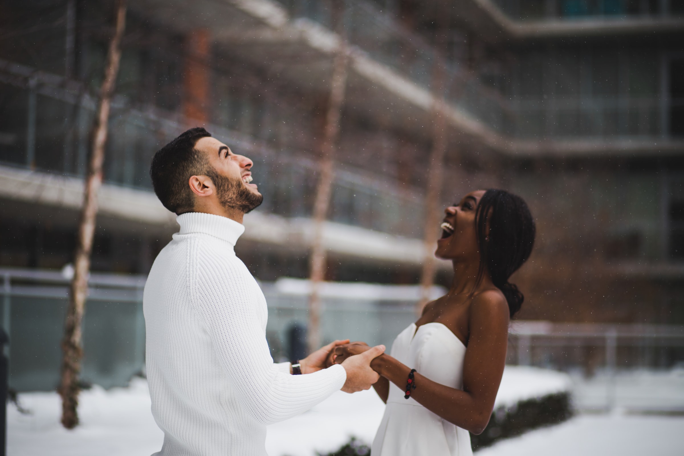 A couple embodying the magicof "Yes, And" by holding hands and smiling up at the sky as it snows.