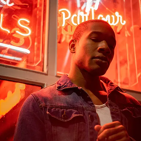 A black man leans against an orange-lit store window with a contemplative face, as if he's wondering if he needs depression counseling.