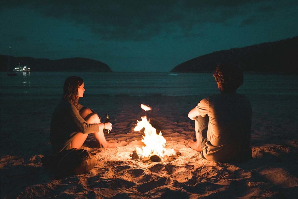 A couple sitting peacefully on the beach by a fire, as if safe from the ugly side of relationship after determining couples therapy is worth it.