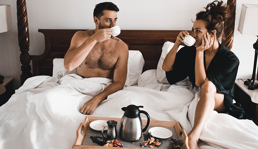 A man and woman in bed eating, resting on their honeymoon.