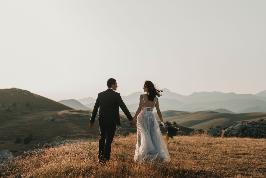 A man holds a woman's hand on a scenic hill, after their wedding, headed to their honeymoon.