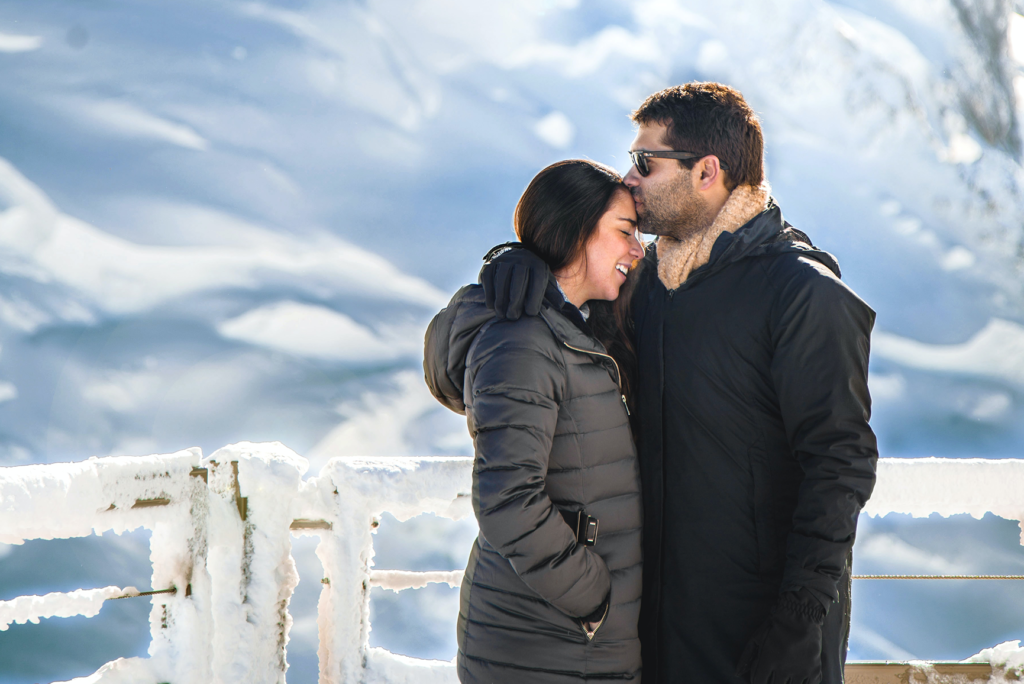A husband kissing his wife's forehead by an icy mountain, doing a honeymoon activity together.