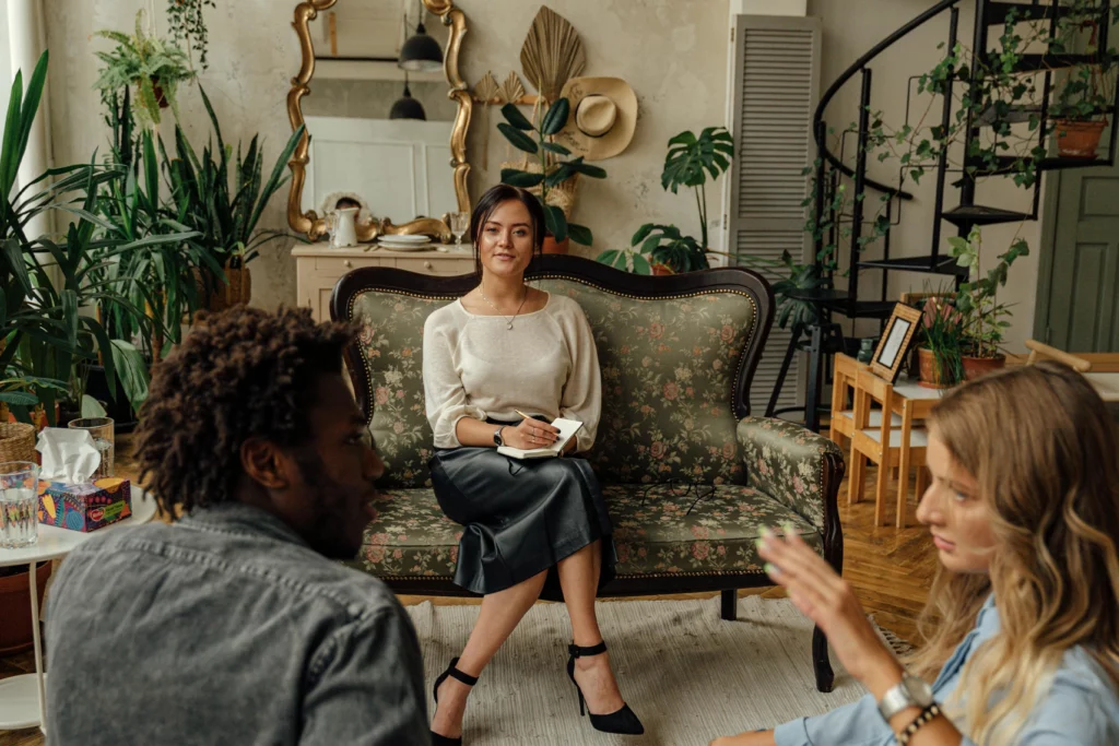 Couples therapist sitting on a couch in a warm, bright, eclectic room, calmly looking at a couple talking in the foreground.