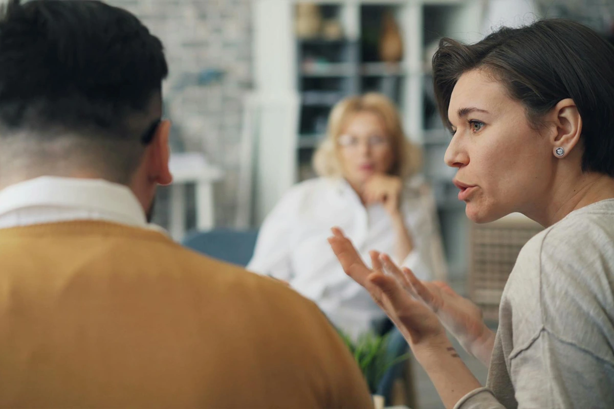 A woman talking, agitated, to her male partner while a couples therapist watches in the background. They could have used our How to Find a Couples Therapist FAQ.