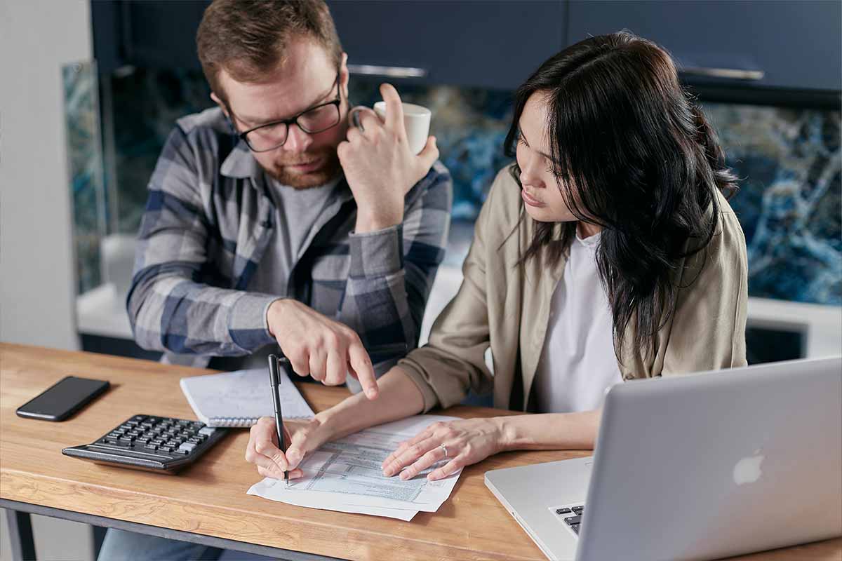 A man and a woman in a relationship sitting at a desk writing concentrating on a printed spreadsheet.