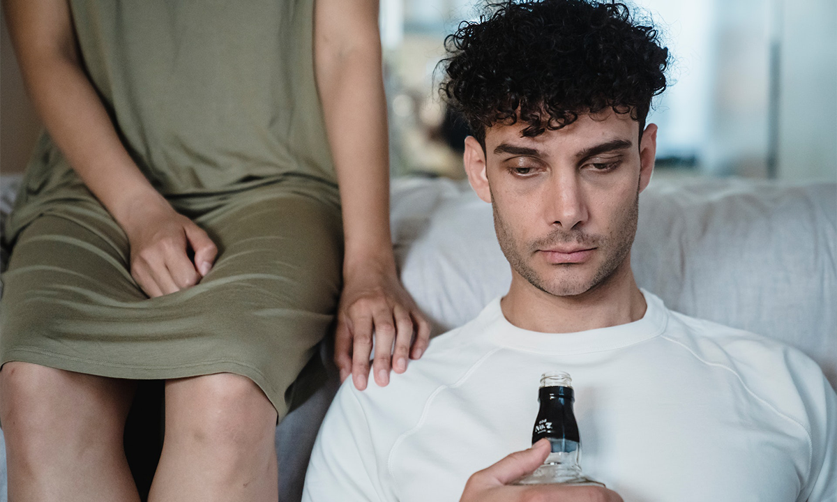 A man sits next to a bed with a bottle in his hand, looking unhappy. His partner, a woman, rests her hand on his shoulder, indicating a relationship with addiction or alcohol and enabling.