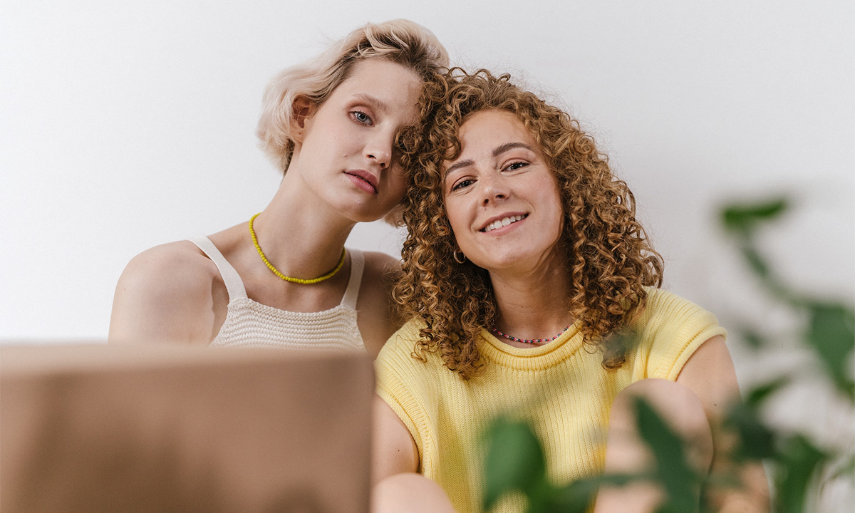 A couple, two women, rest their heads together, looking at the camera. One girl is smiling, the other is serious, as if she's going along with the relationship without meeting her needs.