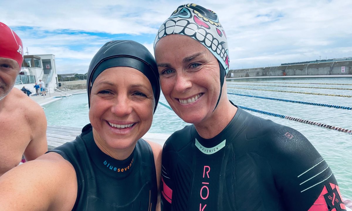 Teale and Amanda smiling for a picture by the pool, wearing wetsuits, with Figs cut off in the background (not wearing a wetsuit).