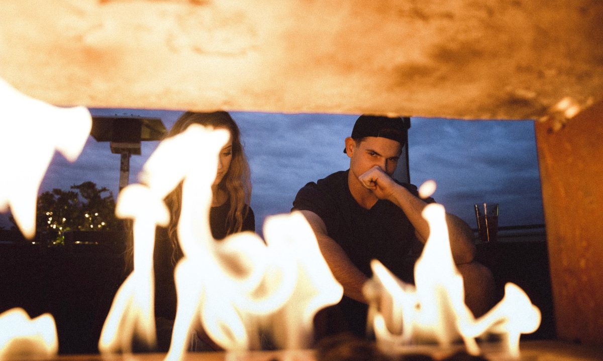A fire from a fireplace blocks the face of a woman, as the man sitting next to her stares into the fire.